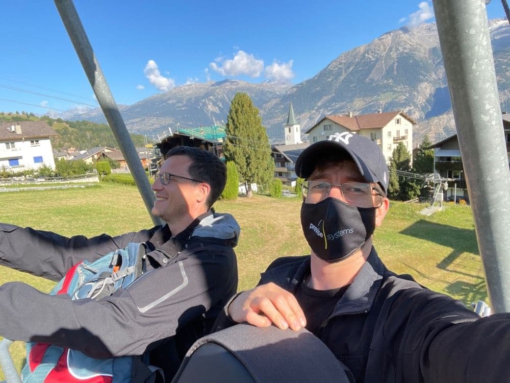 Two people on a ski lift, one wearing a mask, with a scenic mountain village and clear blue sky in the background.