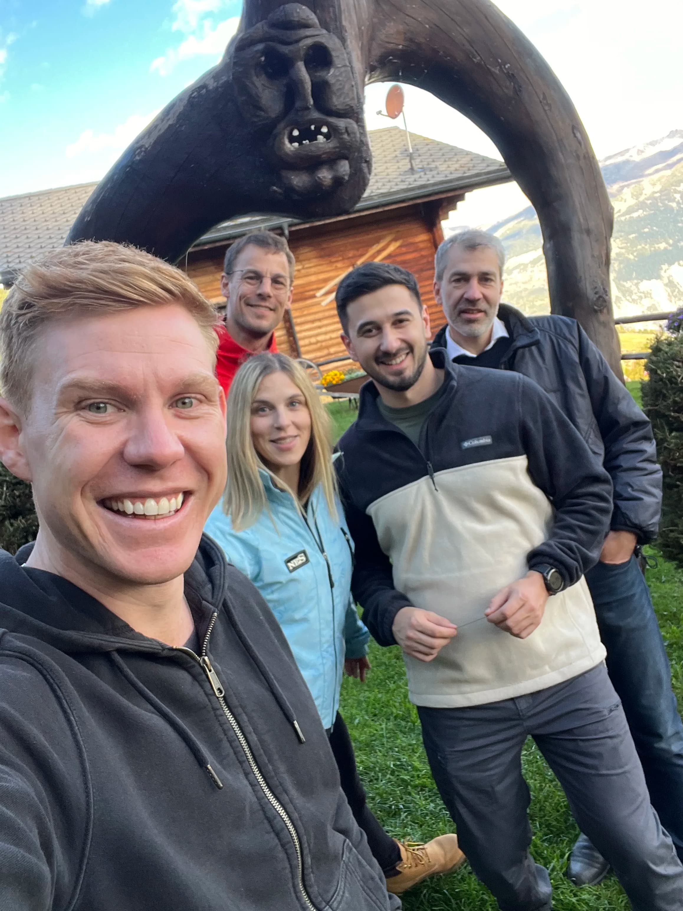 Five people smiling outdoors in front of a wooden sculpture with a cabin and mountains in the background.