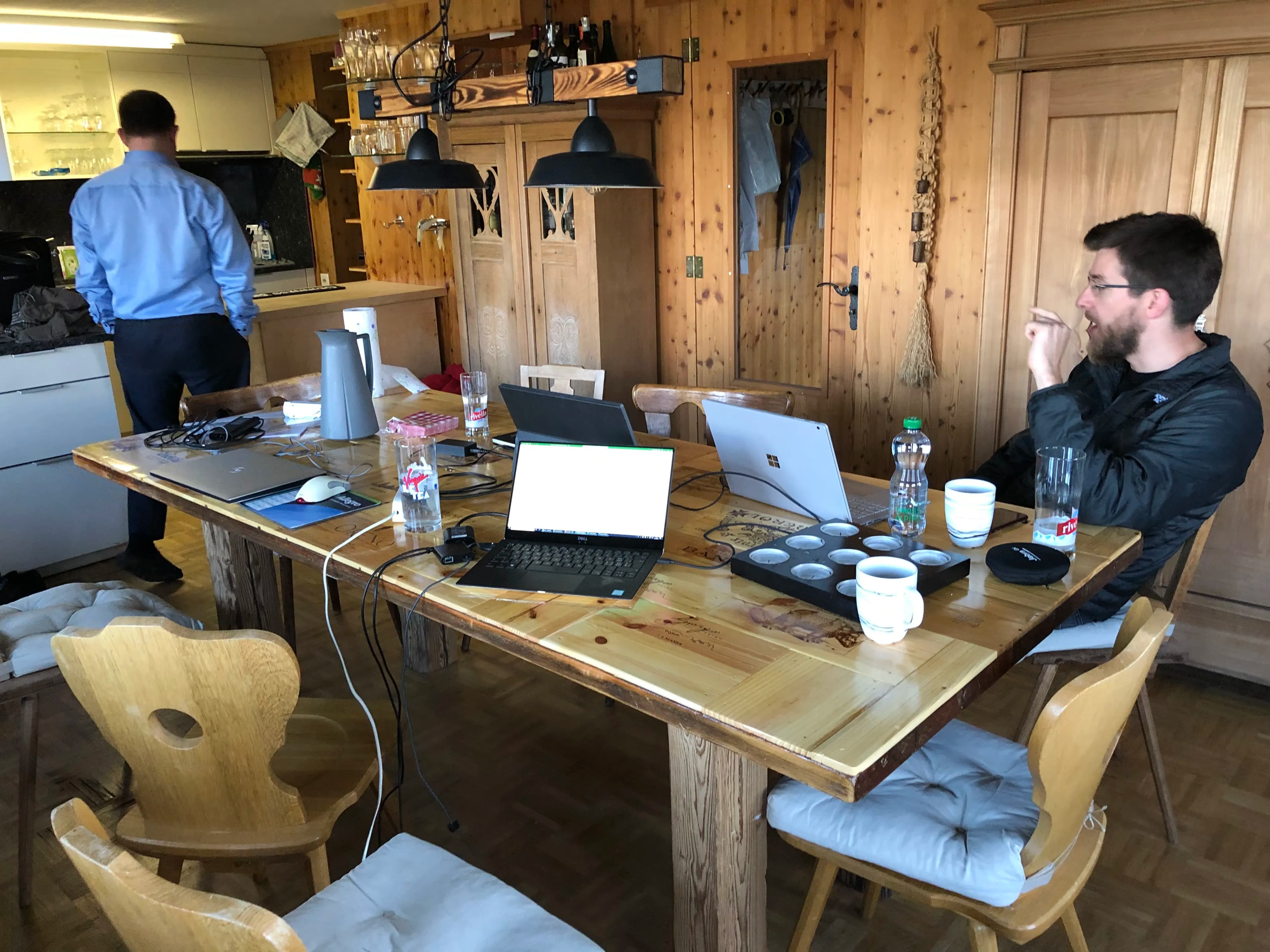 A wooden dining table with laptops, mugs, and bottles. Two men are present, one standing in the kitchen and the other sitting at the table.