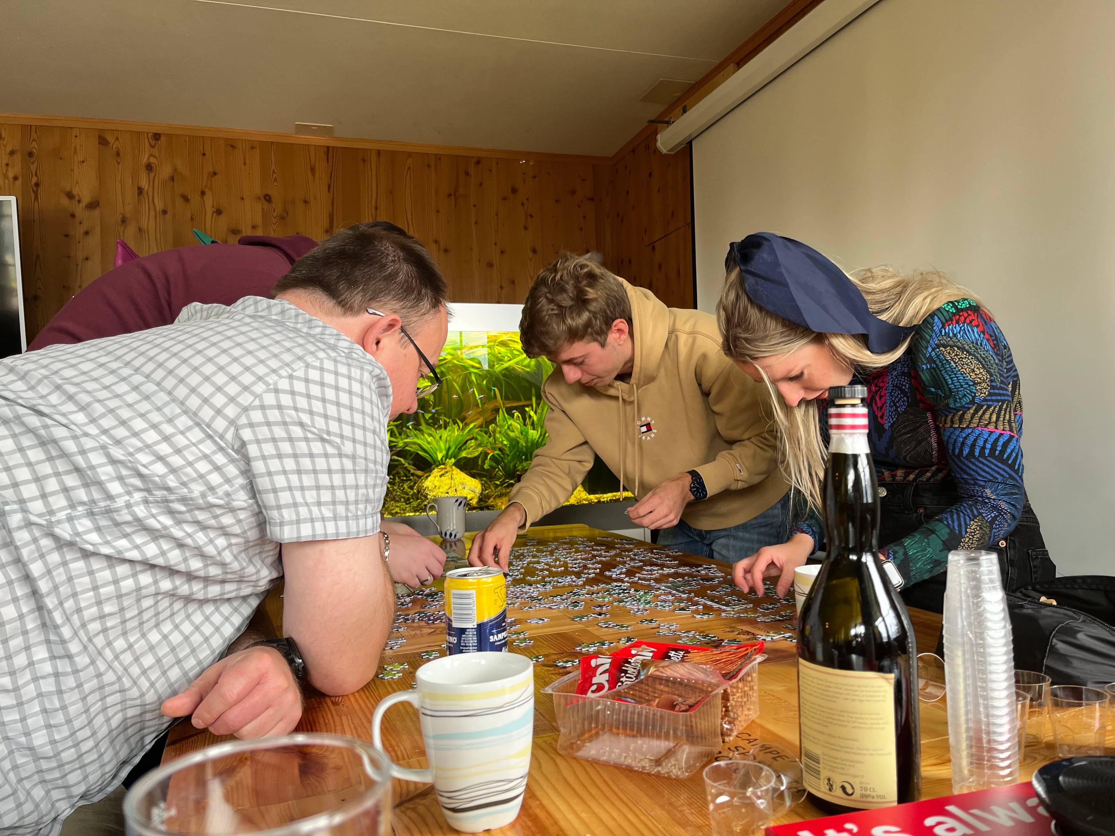 Four people are gathered around a table working on a jigsaw puzzle. Drinks and snacks are visible on the table.
