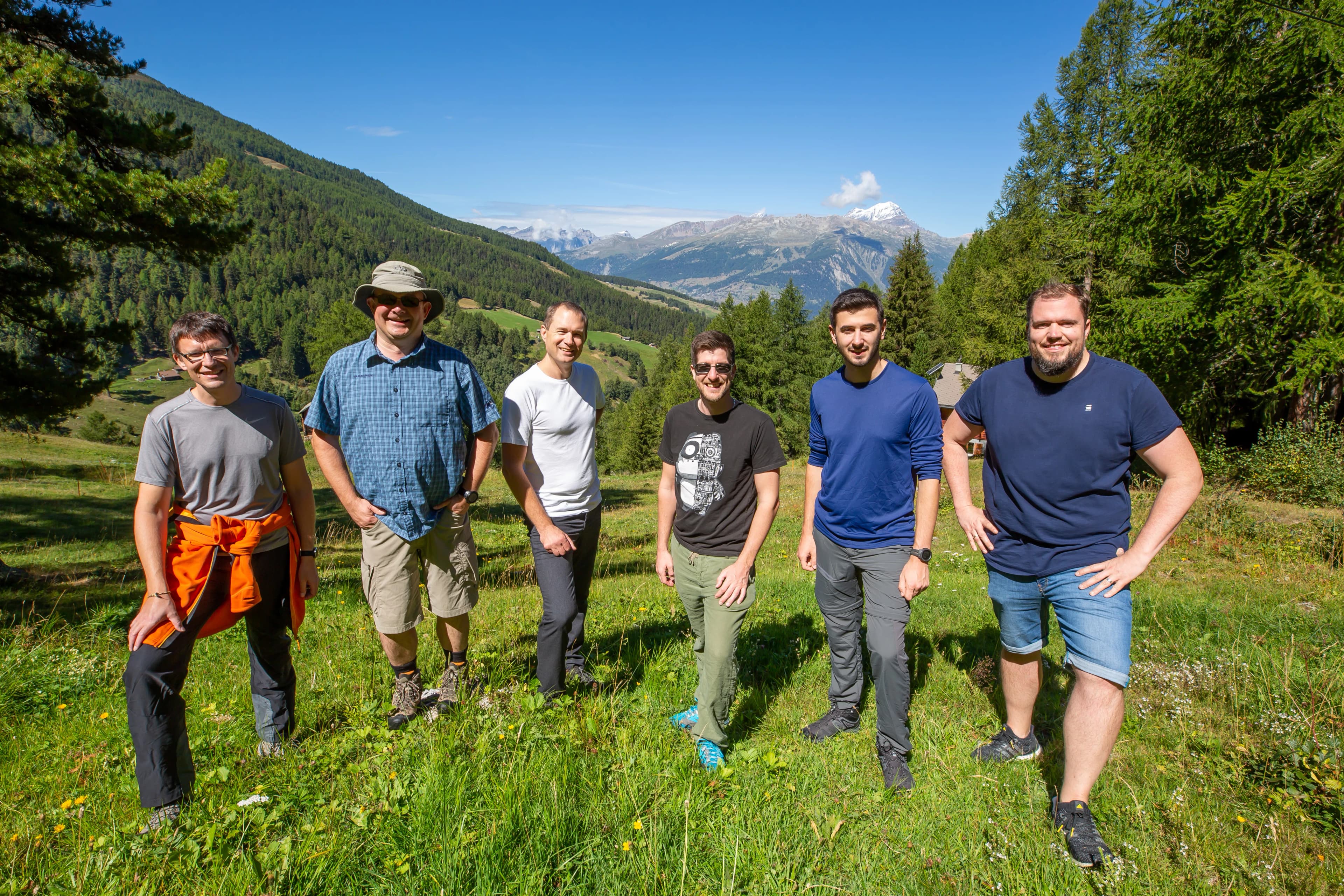 Previse people standing on a grassy hillside with mountains in the background, smiling under a clear blue sky.
